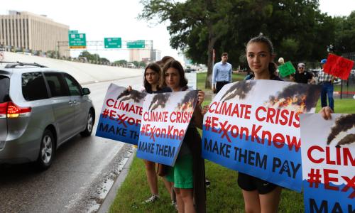 Young girls on the side of the road hold signs that read "617888九五至尊娱乐 Crisis, #ExxonKnew Make Them pay."