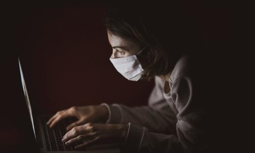 Person wearing a mask in the dark, typing on a laptop.