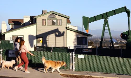 Two people and their dog walking past a house. A pumpjack is in the background.